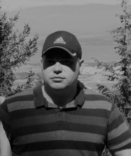 Pic of me above the waist, White male with a striped collared shirt, wearing a ballcap framed by two trees with a valley and hills in the background